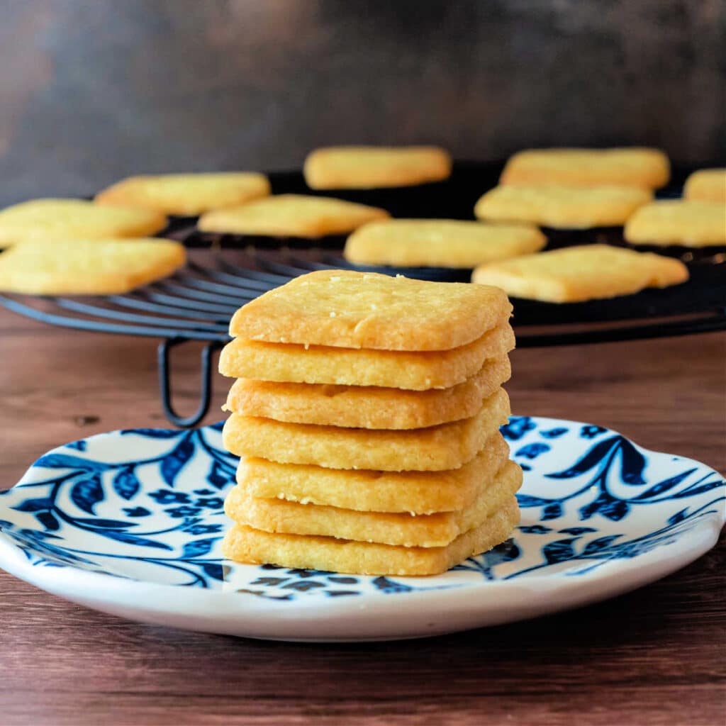 A stack of thin shortbread cookies on a blue plate with a wire rack of more cookies cooling in the background.