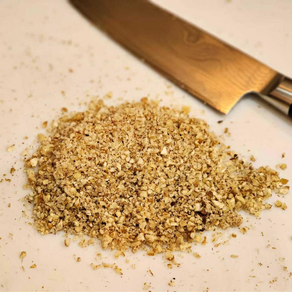 A pile of very finely chopped nuts on a white cutting board with a chef knife in partial view