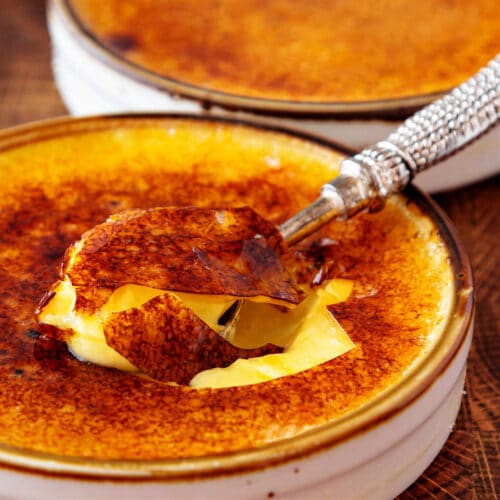 A square close up of a torched creme brulee with a small, silver spoon in it. The sugar is deeply caramelized and very thin with a couple of shards sticking up.