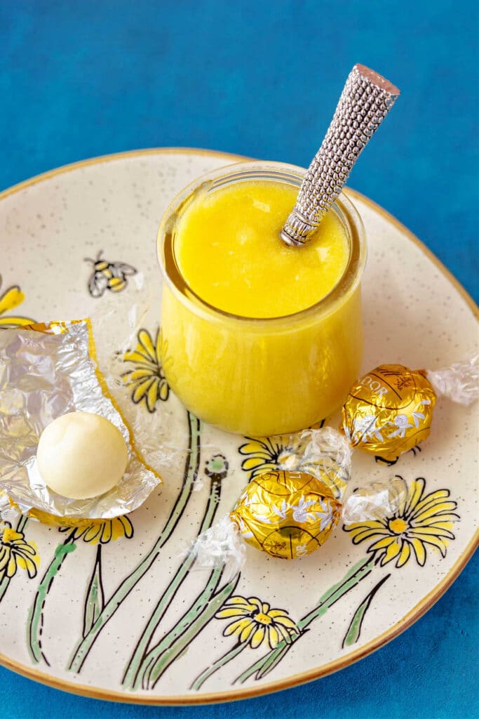 A glass jar of lemon curd with a spoon in it on a plate with a daisy design and white chocolate truffles on it.