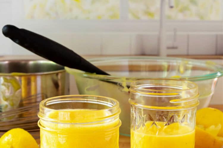 Two glass jars, one half-full and one full, of lemon curd on a butcher block surface with a pan and a glass bowl in the background.