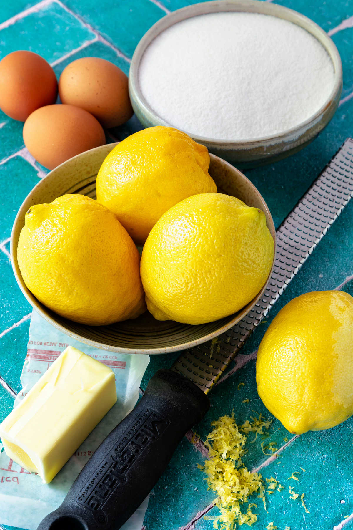 A bowl with 3 lemons in it, a lemon partially zested next to a Microplane with a black handle, a half stick of butter, 3 eggs, and a bowl of sugar.