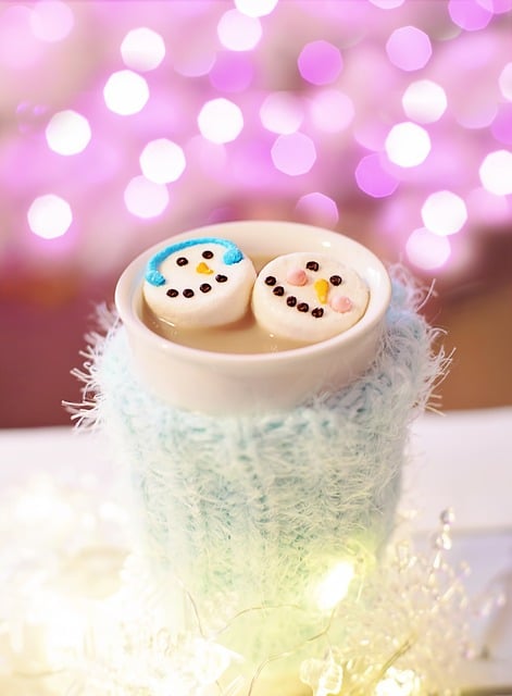 A white mug with a fuzzy light blue mug holder. Marshmallows decorated as snowmen float on top of cocoa. Pastel twinkle lights are in the background and white snowflake lights are in the foreground.