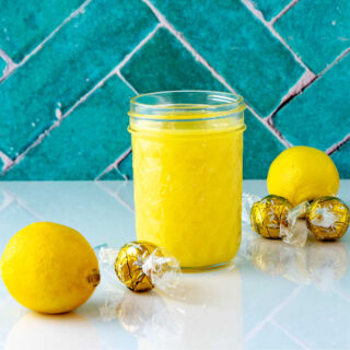 A square image of a glass jar of lemon curd, two lemons, and three wrapped white chocolate truffles on a white reflective surface with a jad tile background.