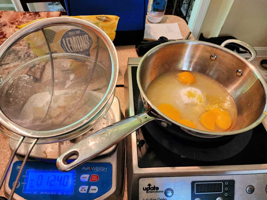 A kitchen scale next to a square induction burner. On the scale is a glass bowl with a strainer set on top. On the induction burner is a metal pan of lemon juice, eggs, egg yolks, and sugar.