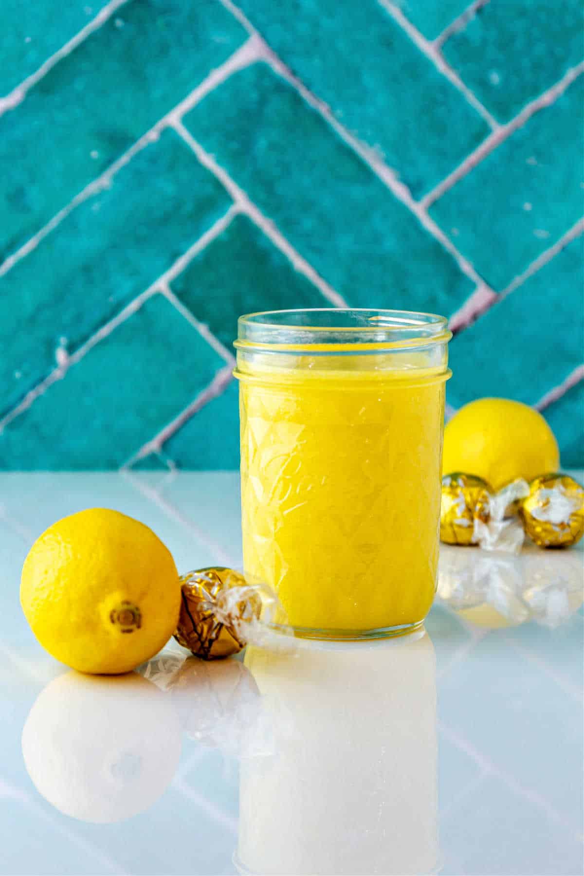 A clear glass jar of lemon curd on a white reflective surface with white chocolate truffles wrapped in foil and lemons.