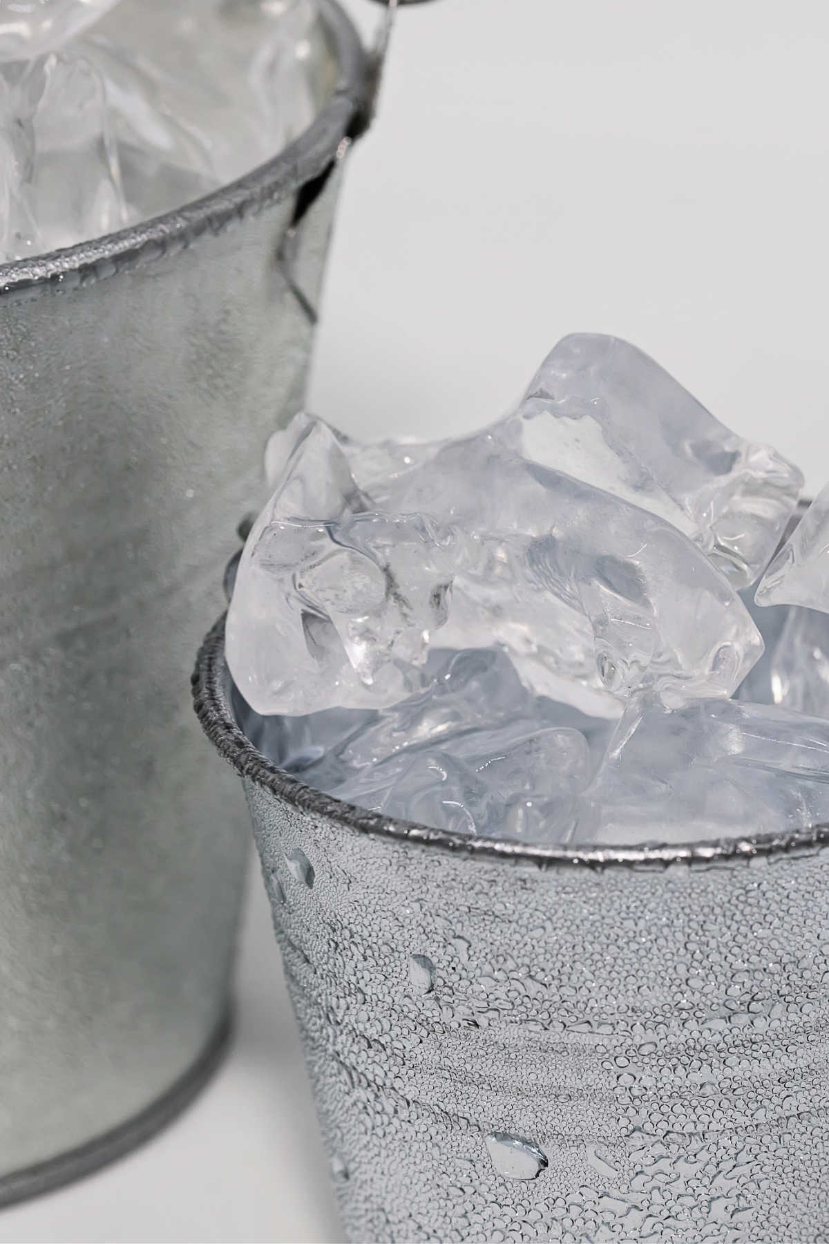 A vertical image of a small metal bucke filled with ice next to a larger bucket of ice.