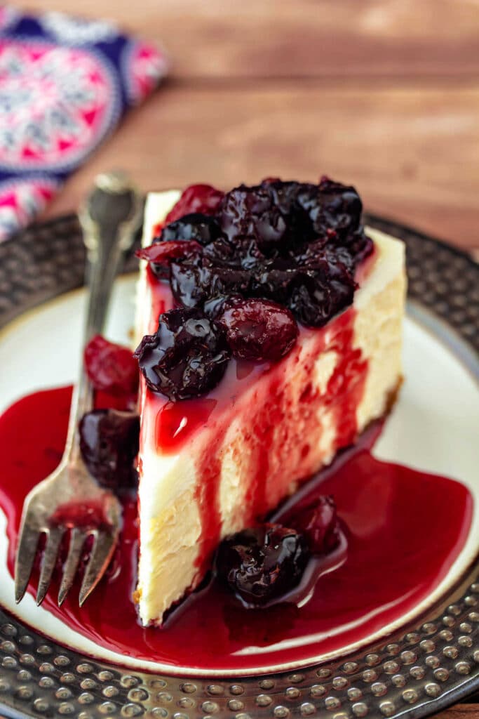 A slice of cheesecake topped with cherry preserves on a white plate with a dark brown border.