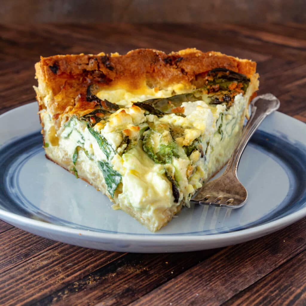 A large slice of quiche with spinach and goat cheese on a blue plate with a fork.