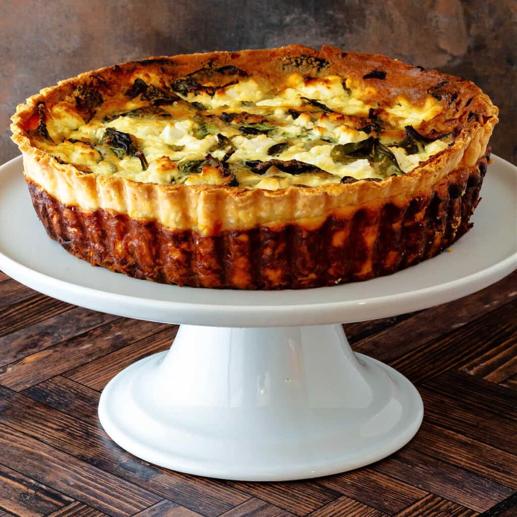 A whole quiche baked in a fluted, deep dish tart pan on a white cake stand.