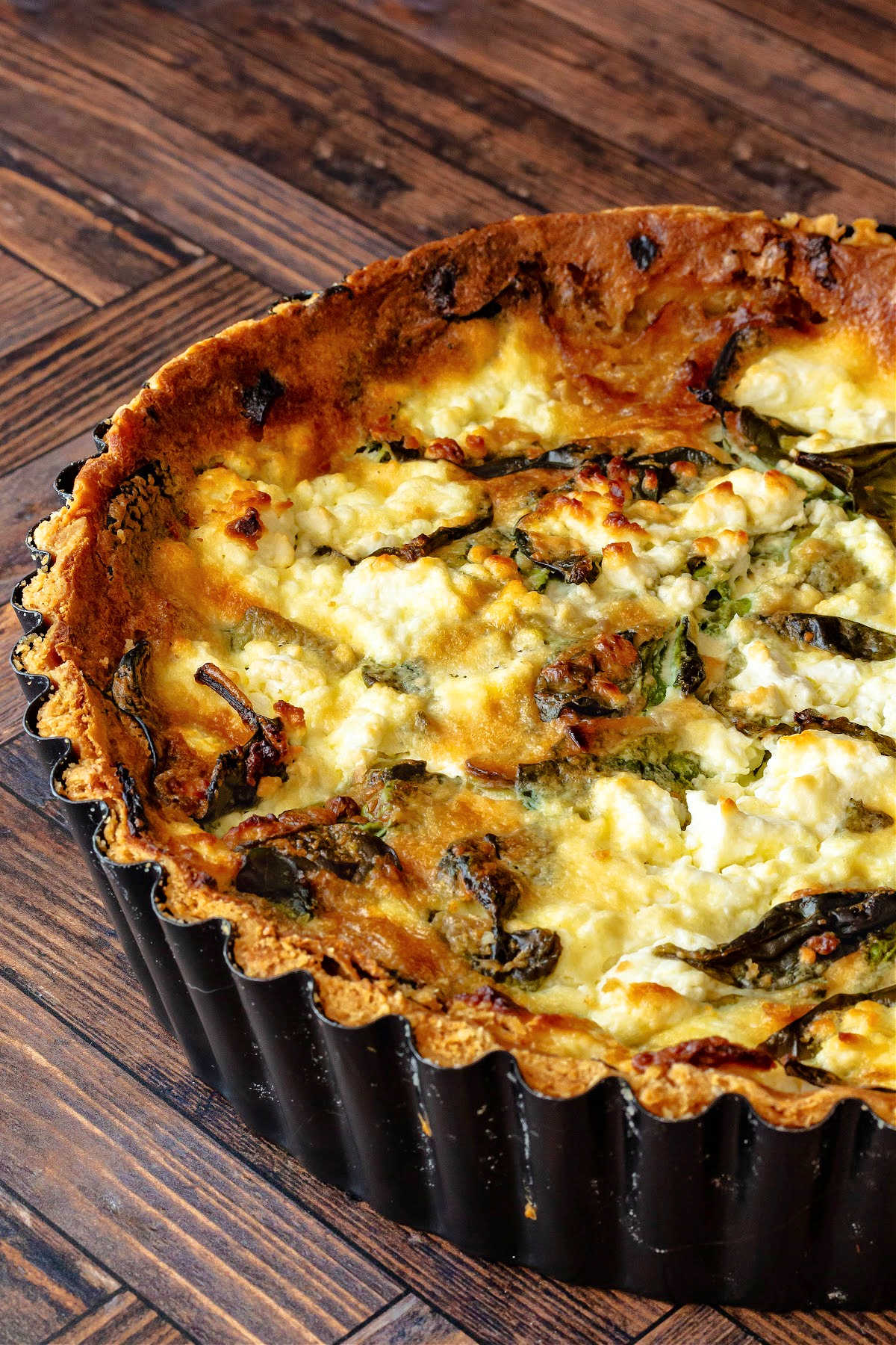 A whole quiche in a black, deep dish tart pan, fully baked. The top is deep golden brown and you can see spinach leaves and goat cheese peeking out of the custard.