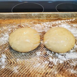 Two balls of pizza dough covered with plastic wrap on a half-sheet pan.