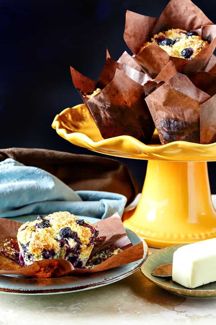 An orange cake stand piled with blueberry muffins in brown paper wrappers with one muffin on a plate.