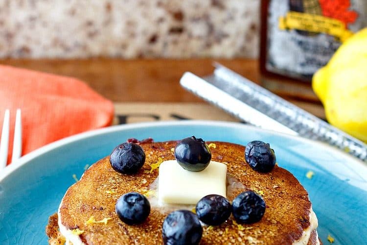 A plate of pancakes with butter and blueberries on top. A bottle of maple syrup is in the background.