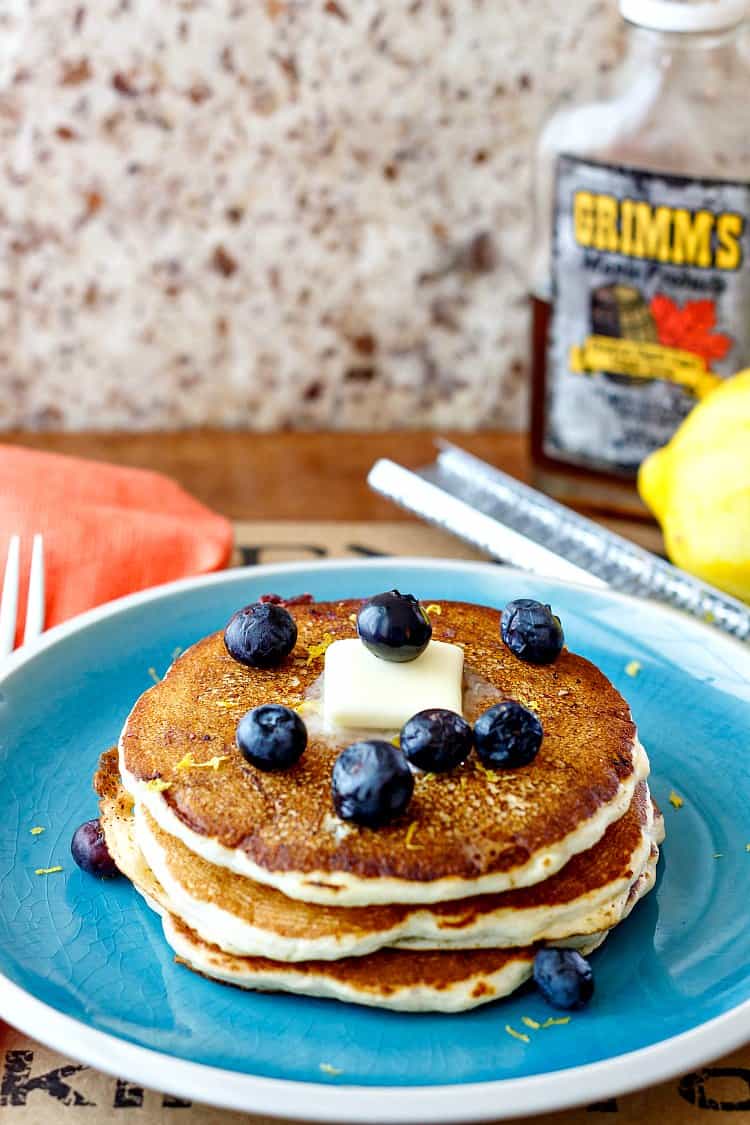 A plate of pancakes with butter and blueberries on top. A bottle of maple syrup is in the background.