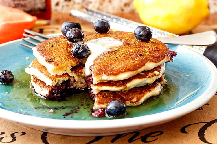 A stack of 3 pancakes with a bite cut out. Melted butter, syrup and fresh blueberries on top.