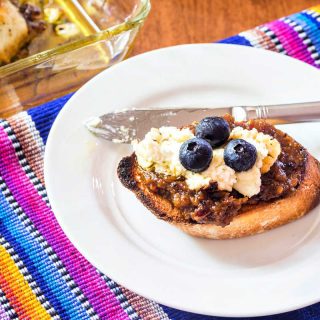 One piece of toasted bread brushed with olive oil and topped with bacon jam, goat cheese, and 3 blueberries on a white plate.