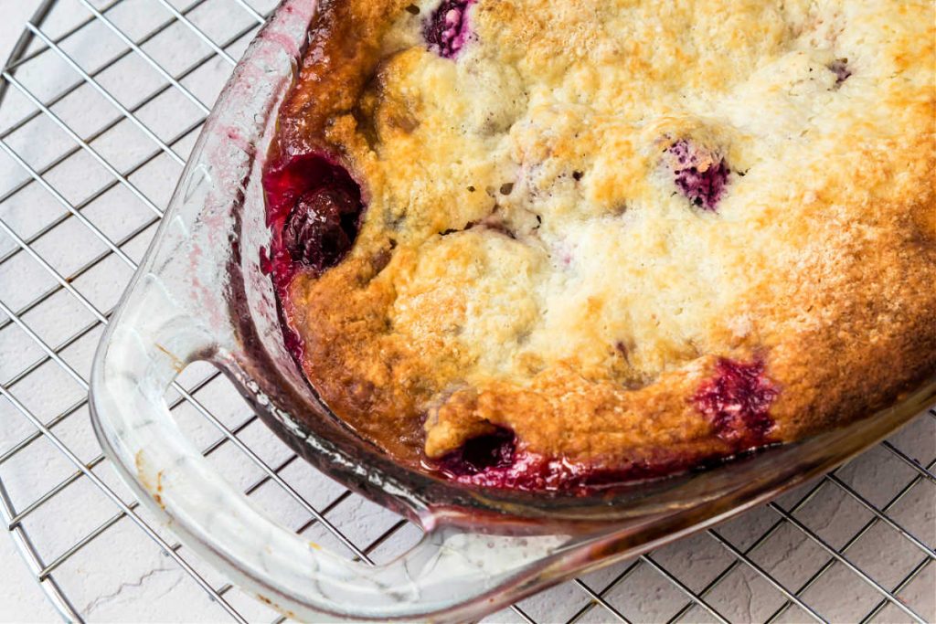 A partial shot of a glass dish of cherry cobbler on a cooling rack. You can see some cherries peeking through the topping.