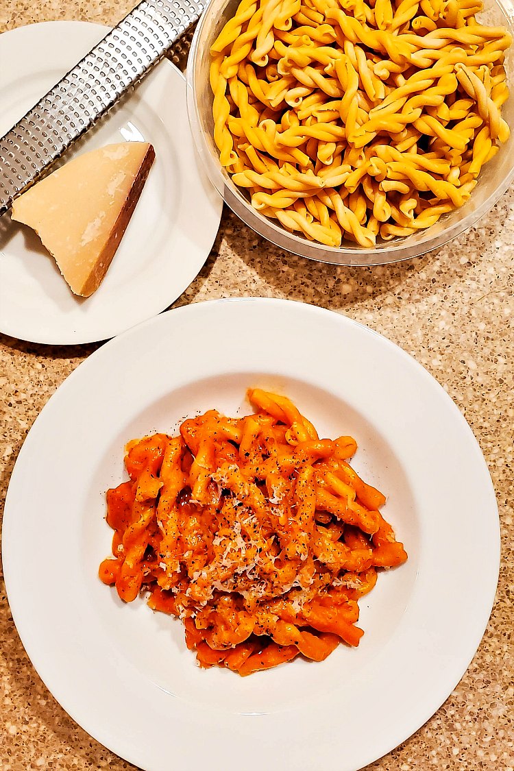 Overhead shot of pasta with sauce on a white plate with a wedge of Parmesan, a microplane grater, and a bowl of dry pasta.