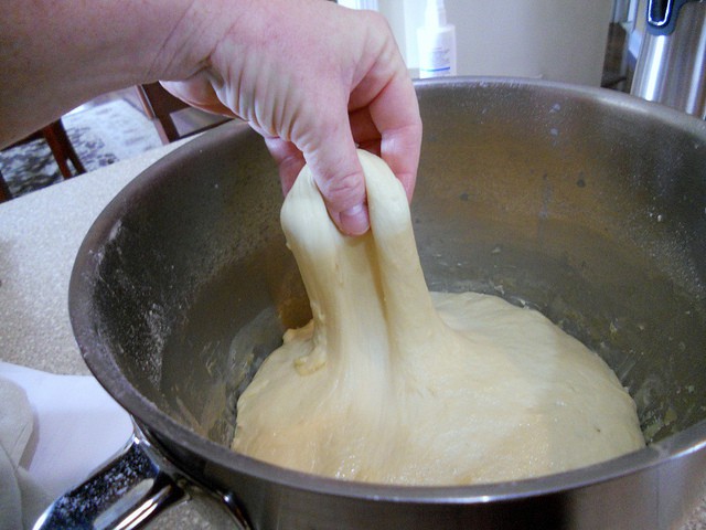 A hand lifting a pinch of sugar cake dough away from main dough, showing the stretch. Dough is in a mixing bowl.