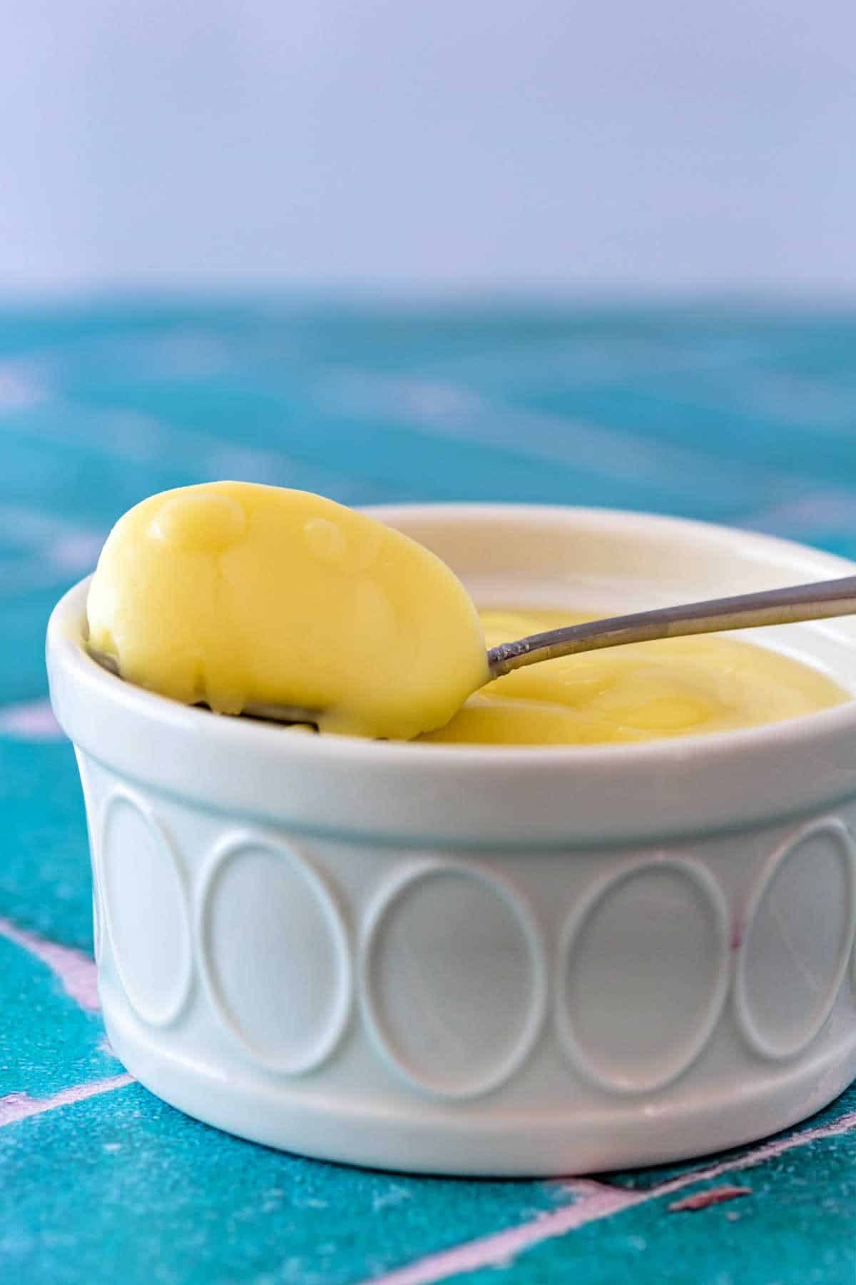 A spoonful of pale yellow lemon posset on a spoon balanced on the edge of a white ramekin of posset.