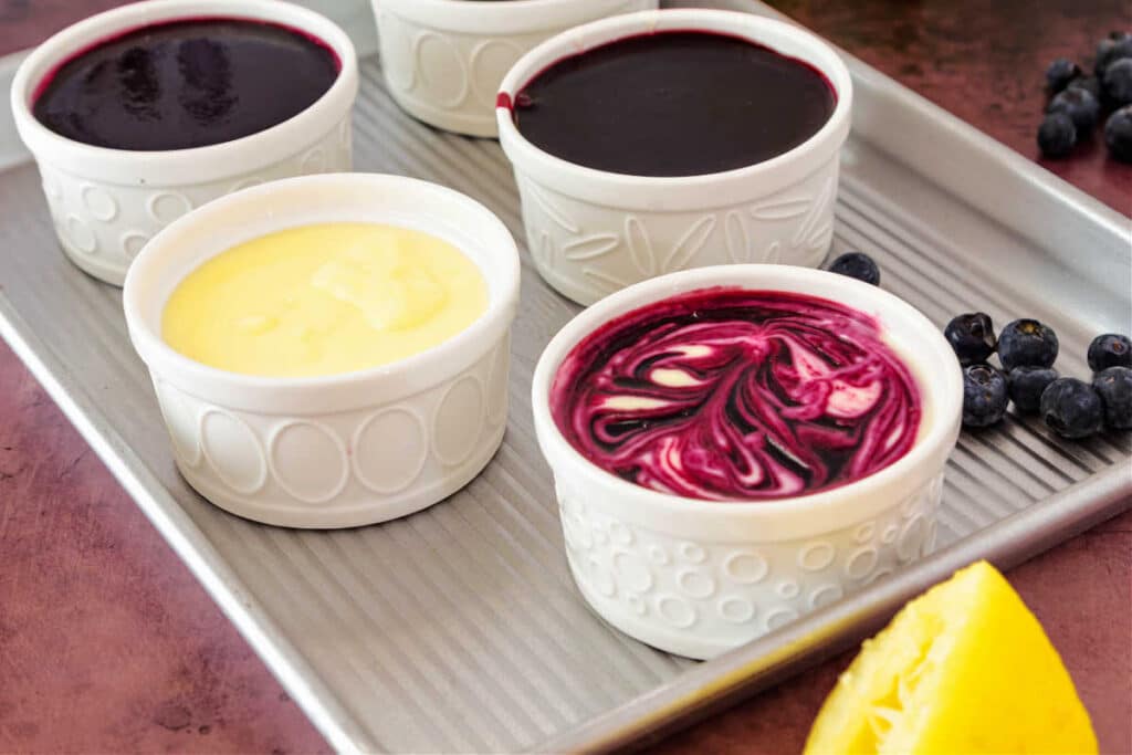 A silver metal tray with white ramekins of dessert on it. One dessert is pale yellow, two are deep purple, and one is yellow and purple swirled.