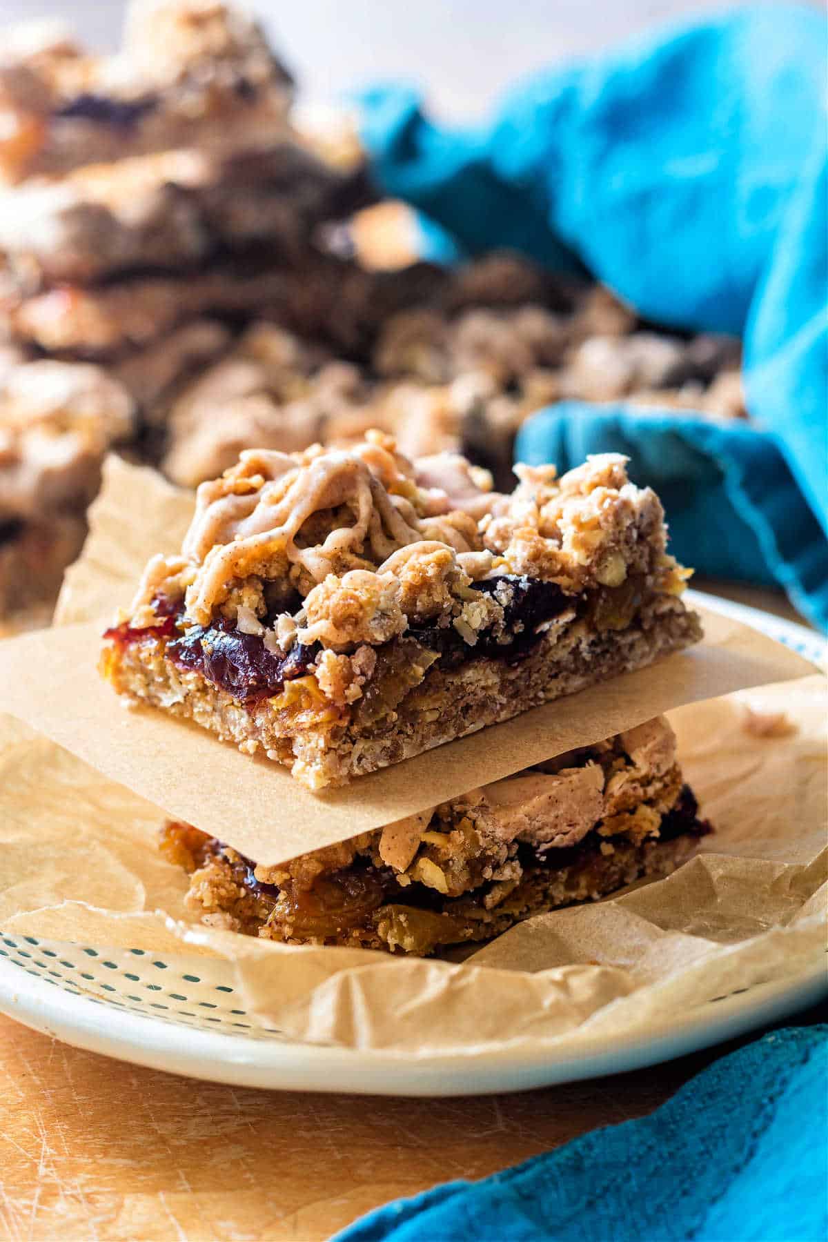 A vertical image of two stacked cranberry raisin bars on a small plate, separated by a small rectangle of parchment. There are more raisin bars in the background as well as a turquoise-colred cloth napkin.