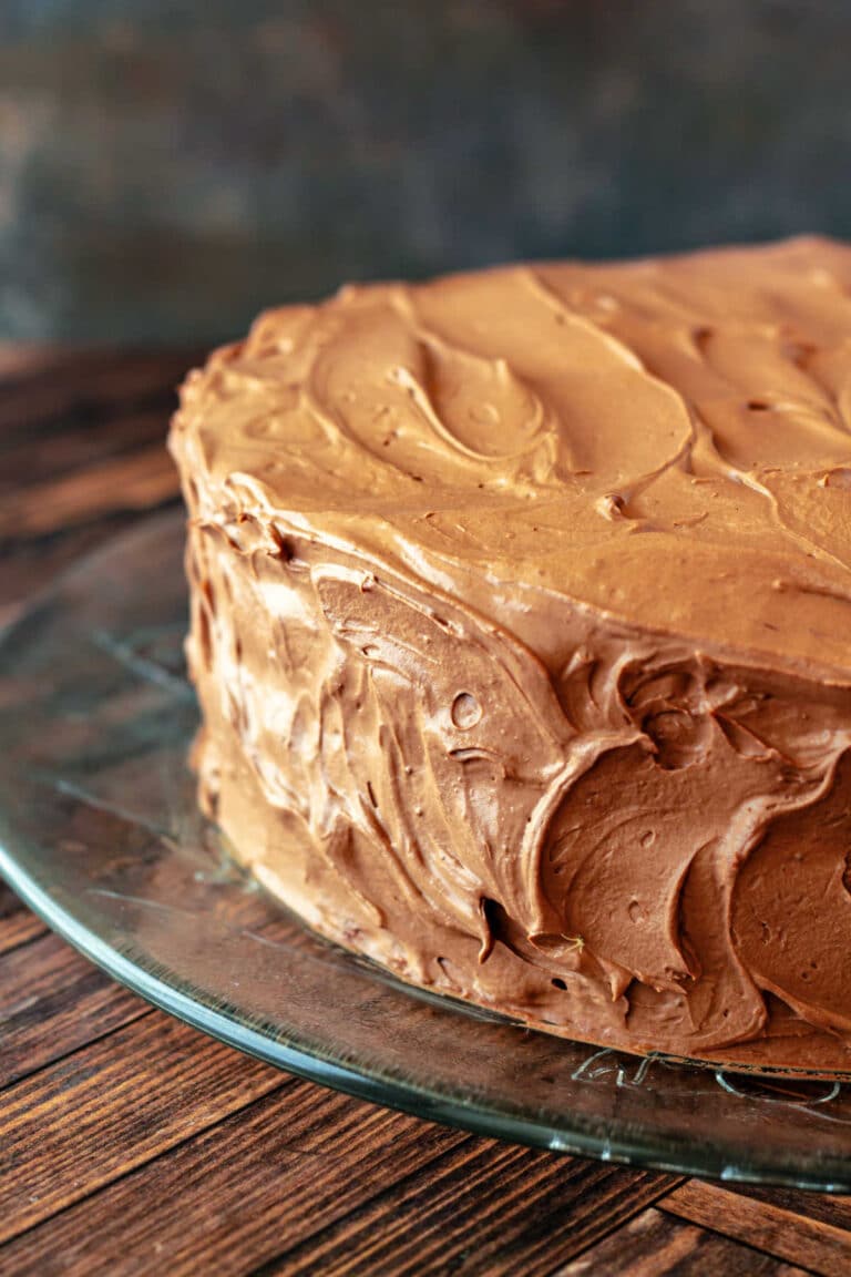 A 3/4 angle partial shot of a whole cake swirled with chocolate frosting on a glass platter.