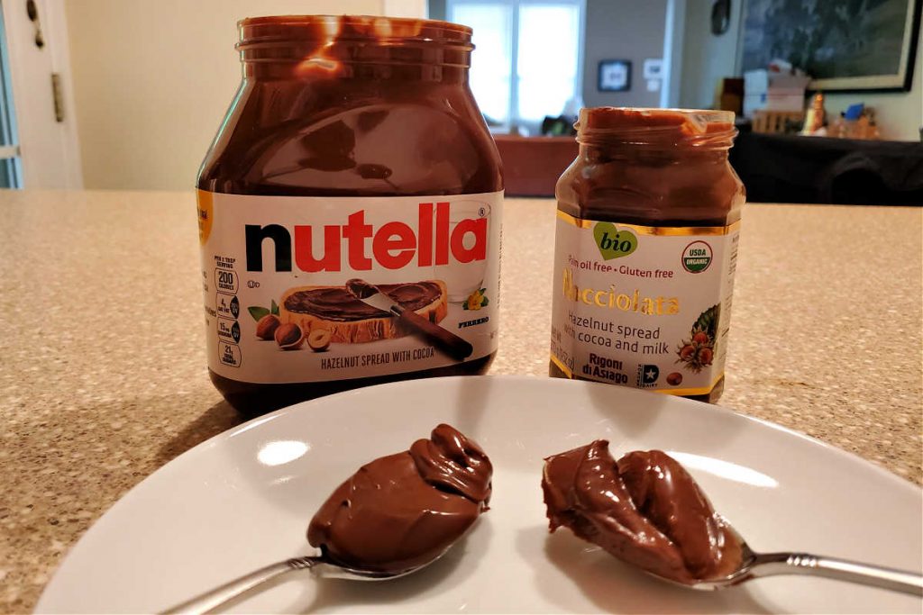 A jar of Nutella and a jar of Nocciolata with spoonfuls of each in front.