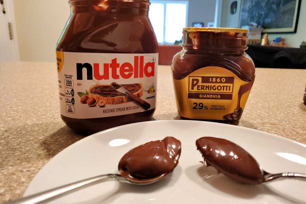 A jar of Nutella and a jar of Pernigotti with spoonfuls of each in front.