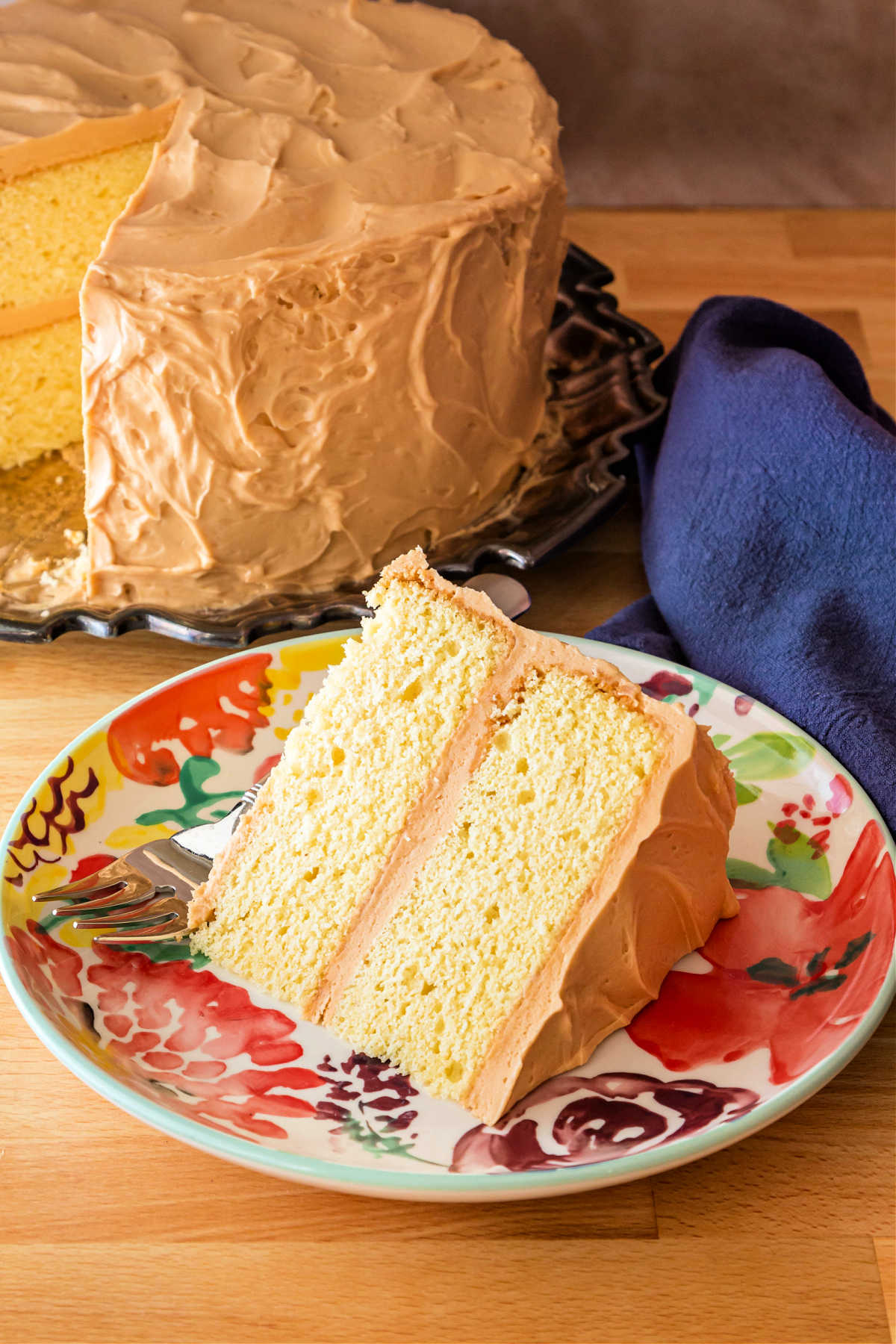 A floral plate with a slice of yellow cake on it with the rest of the cake in the background.