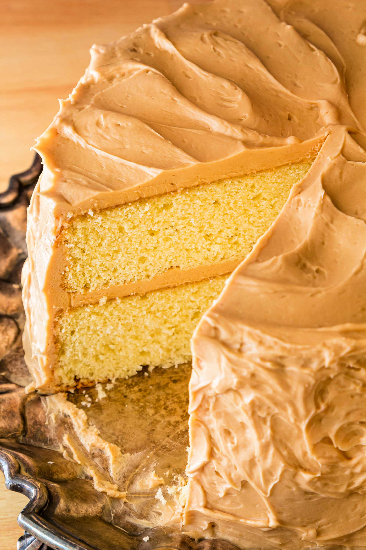 A 3/4 angle view of a whole 1-2-3-4 yellow cake frosted with beige frosting with one large slice cut out of it. Two layers of yellow cake with beige frosting in between are visible on the cut side of the cake.