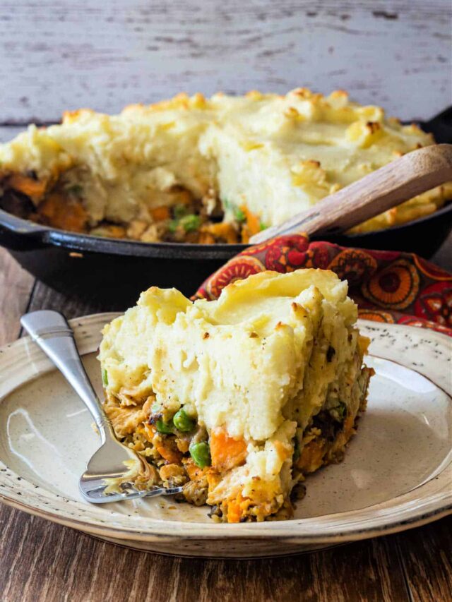A cut slice of savory turkey pie topped with mashed potatoes on a plate with the cast iron skillet of shepherd's pie in the background.