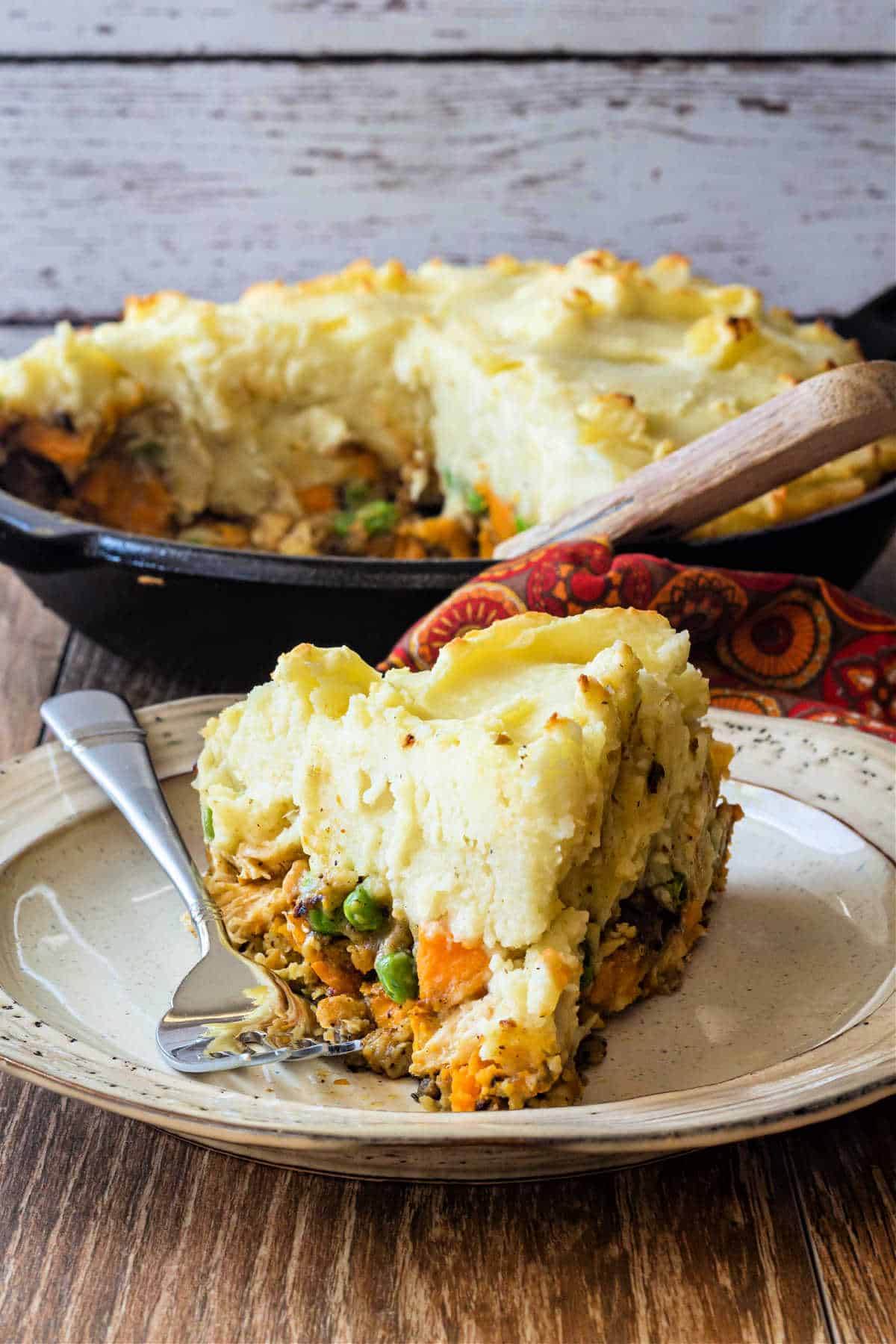 A cut slice of savory turkey pie topped with mashed potatoes on a plate with the cast iron skillet of shepherd's pie in the background.