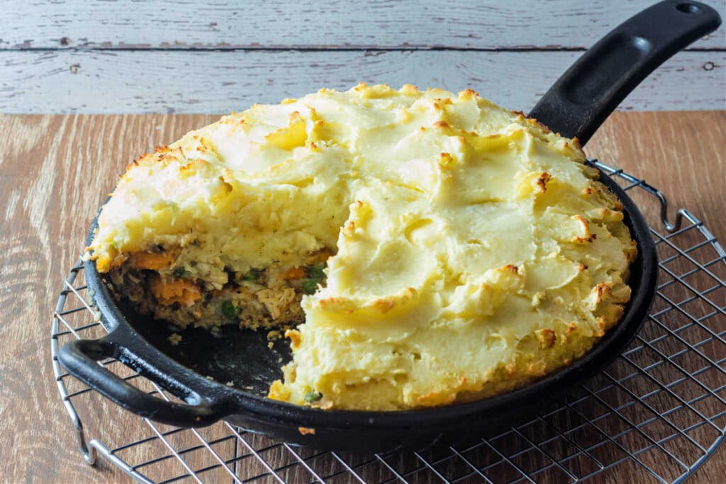 A cast iron skillet filled with thanksgiving shepherd's pie with a slice cut out of it. The pie has a crust made out of stuffing, is filled with turkey, sweet potatoes, peas, cranberries, and gravy and is topped with mashed potatoes.