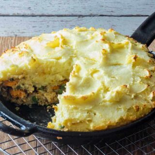 A cast iron skillet filled with thanksgiving shepherd's pie with a slice cut out of it. The pie has a crust made out of stuffing, is filled with turkey, sweet potatoes, peas, cranberries, and gravy and is topped with mashed potatoes.