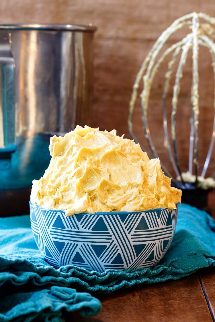 A mound of the best caramel buttercream frosting in a blue bowl with the mixer bowl and whisk in the background.