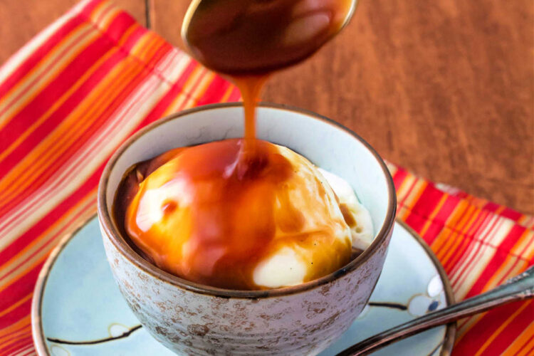 A spoon of caramel sauce dripping down over vanilla ice cream in a blue bowl.