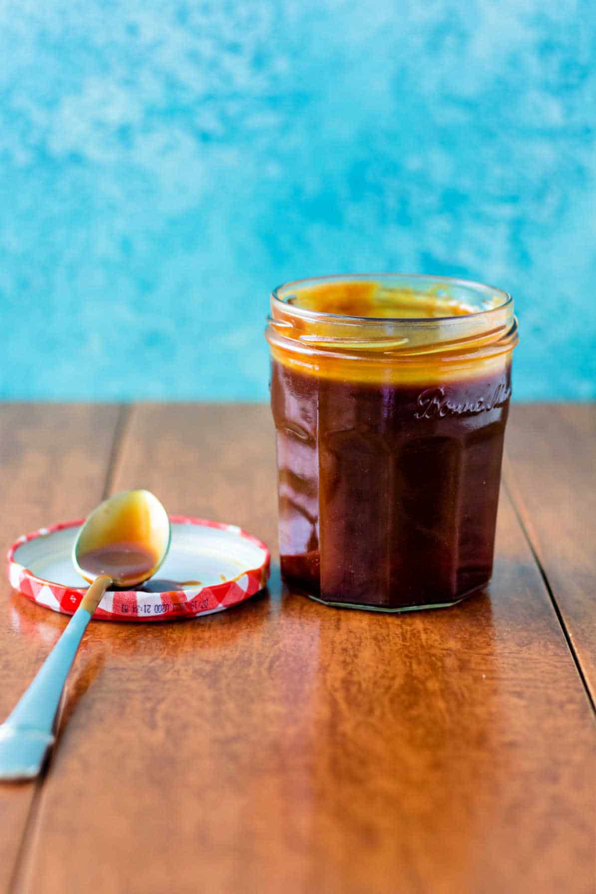 A nearly-full jar of caramel sauce. The lid is off and placed to the left of the jar. A spoon rests on the lid.