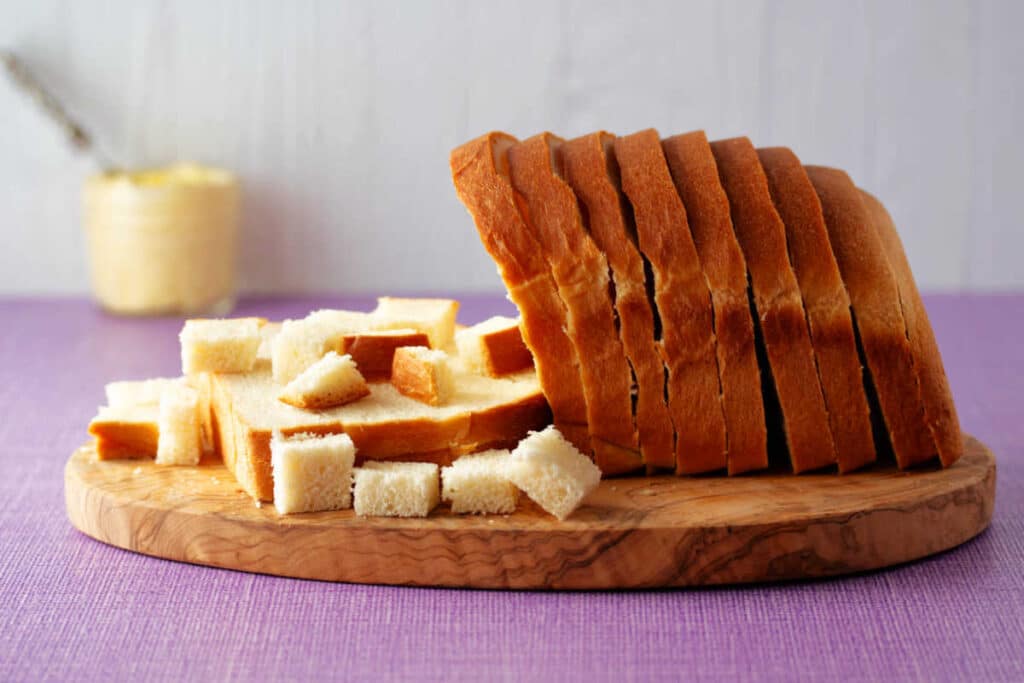 A small cutting board with sliced bread and small squares of bread on a lilac linen surface with a glass jar of whipped butter in the background.