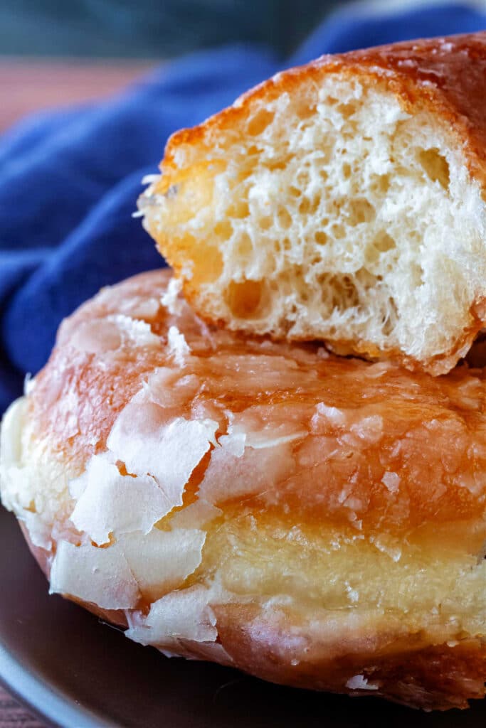 A close-up of part of two donuts stacked on top of each other. The bottom one shows the thin, crackly glaze, and the top one has a bite out of it showing the interior crumb structure.