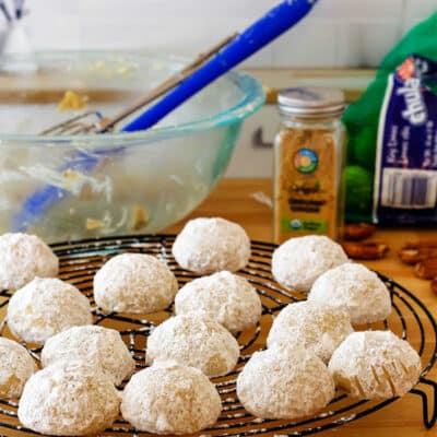 Wedding cookies coated in powdered sugar on a cooling rack with a bowl and spatula in the background.