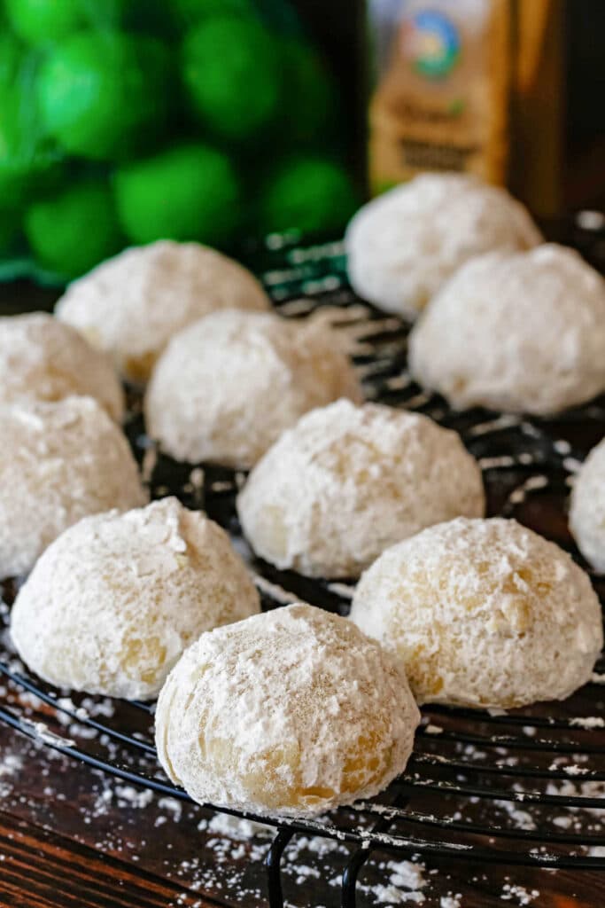 Ginger lime wedding cookies coated in powdered sugar on a cooling rack with key limes and a jar of ginger in the background.