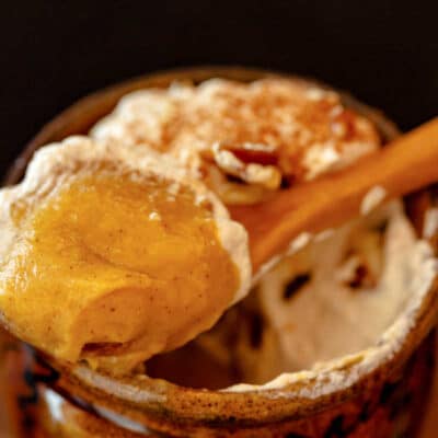 A bite of pumpkin pudding and whipped cream on a small wooden spoon resting on a small ramekin of pudding.