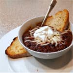 A white bowl of rich, dark chili topped with shredded jack cheese and a dollop of sour cream.