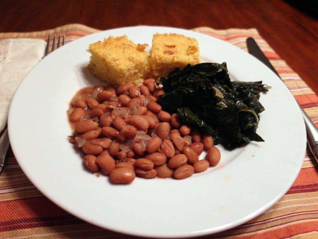 A white dinner plate showing a Poor Man's Dinner of pinto beans, cooked greens and cornbread.