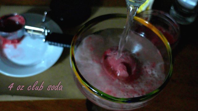 Raspberry Fizz Cocktail being poured into a margarita style glass. Beverage is fizzing and pink raspberry color is prominent in glass. Text in picture says, "4 oz club soda".