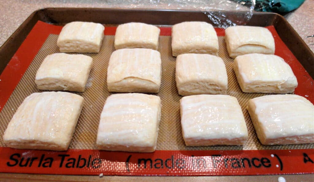Twelve square biscuits on a Silpat before baking. The tops of the biscuits have been brushed with buttermilk.