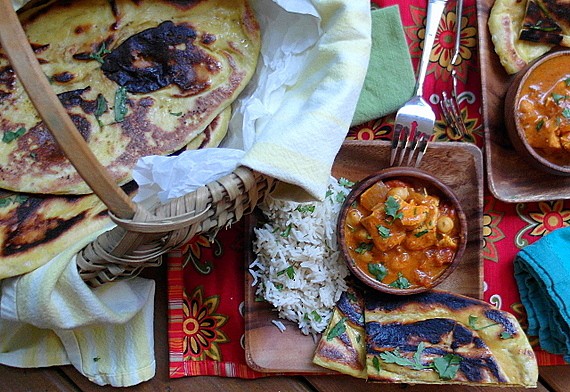 Kashmiri Naan and an Indian meal on a table. Naan loaves are in a rattan basket. Basmati rice is on a square wooden plate. An indivdiual sized serving bowl is filled with a stew that is predominantly red and chunky, topped with chopped fresh herbs. There is a colorful red and yellow placemat under wooden plate. 