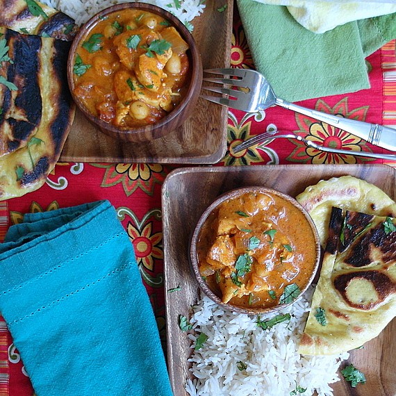 An Indian meal featuring Kashmiri Naan on two wooden plates along with rice and a chunky red stew. All are on a tablecloth of red and yellow flowers. There is a blue napkin next to one plate and a green napkin next to the other plate. 